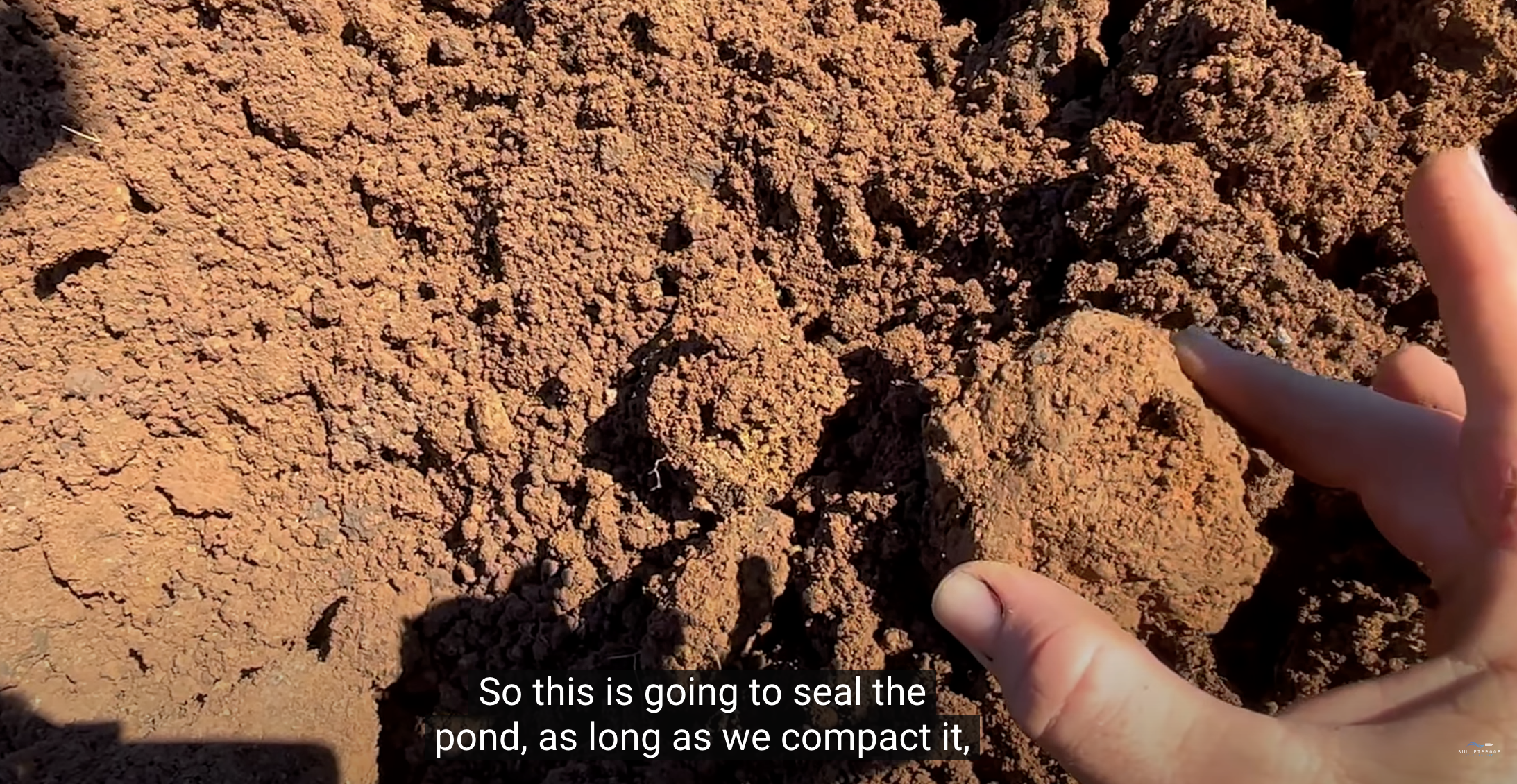Hand checking red clay soil on a pond site; suitable clay only seals a pond when it's properly compacted