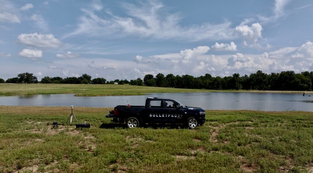 LiDAR Drone Surveying for Ponds and Lakes for Accurate Professional Planning by Bulletproof Pond and Lake with Bulletproof truck and logo and lake construction - Bulletproof Pond and Lake Black pickup truck with the word 'Bulletproof' parked near a calm pond, surveying gear in the foreground on a grassy field under a blue sky.