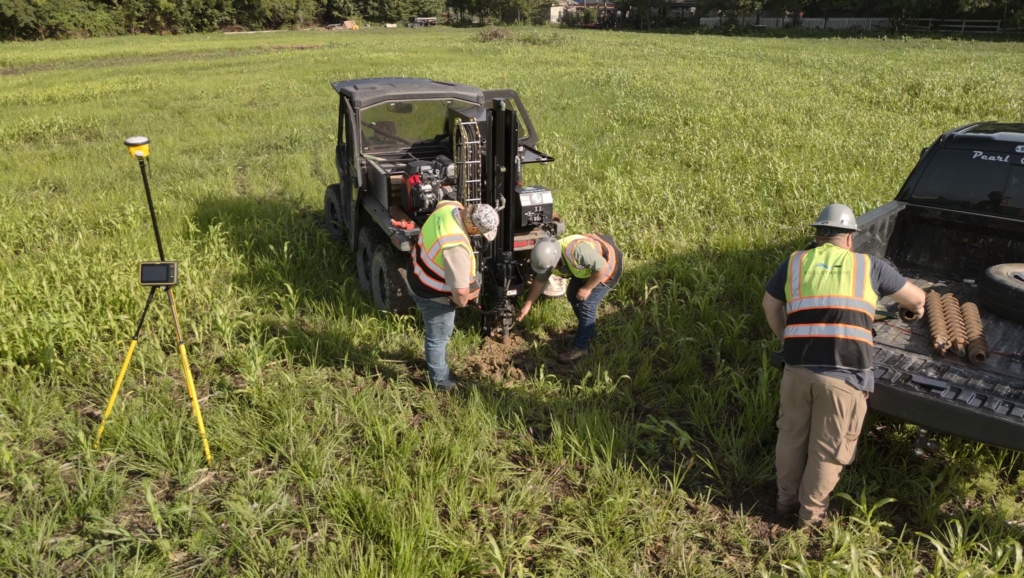 Drill Rig for Soil Borings Professional Pond and Lake Builder Soil Testing Machine by Bulletproof Pond and Lake - Bulletproof Pond and Lake Workers in safety vests operate a soil drill rig in a grassy field beside a pickup truck, with a surveying tripod on the left.