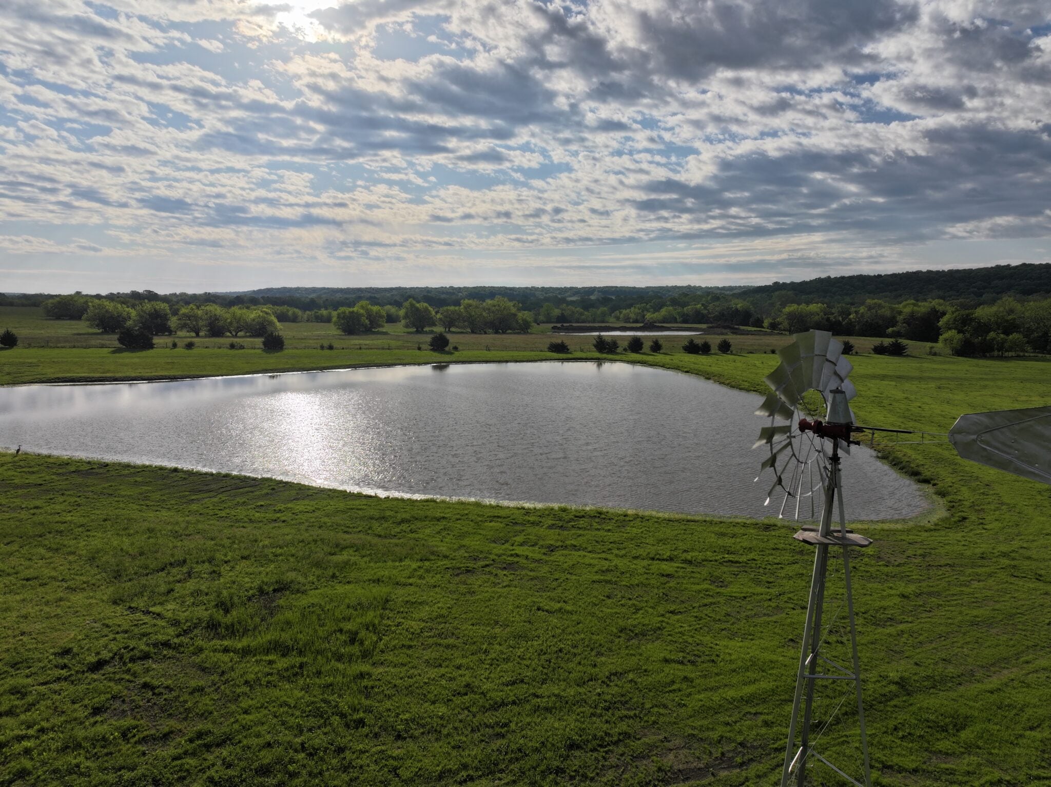 Wide rural landscape with a small pond, green fields, trees, and a metal wind sculpture in the foreground under a cloudy sky.