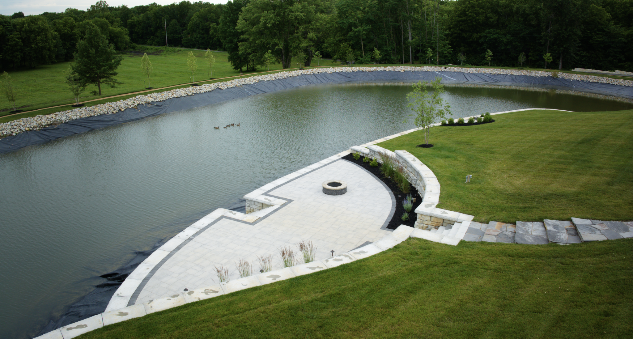 Aerial view of a curved, white stone terrace beside a rectangular pond with a grassy lawn and rock-lined berms along the water's edge.