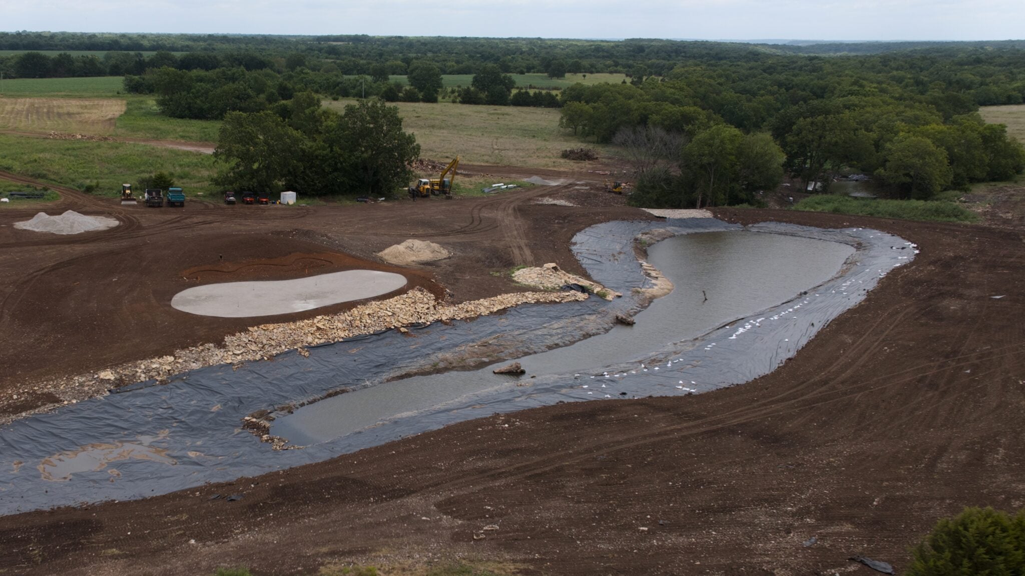 Large excavation site with a winding mud channel and a muddy pond under construction, surrounded by trees and dirt roads