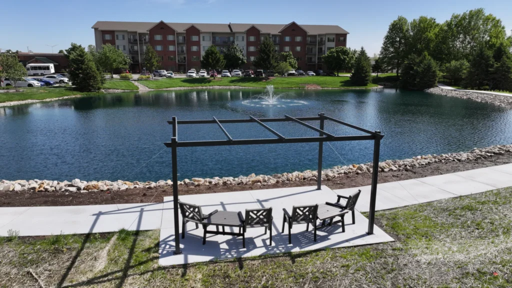 Pond Makeover with concrete walkway, entertaining space, and treed landscaping with fountain, bridge, and fish feeder in Olathe Kansas. Completed by Bulletproof Pond and Lake