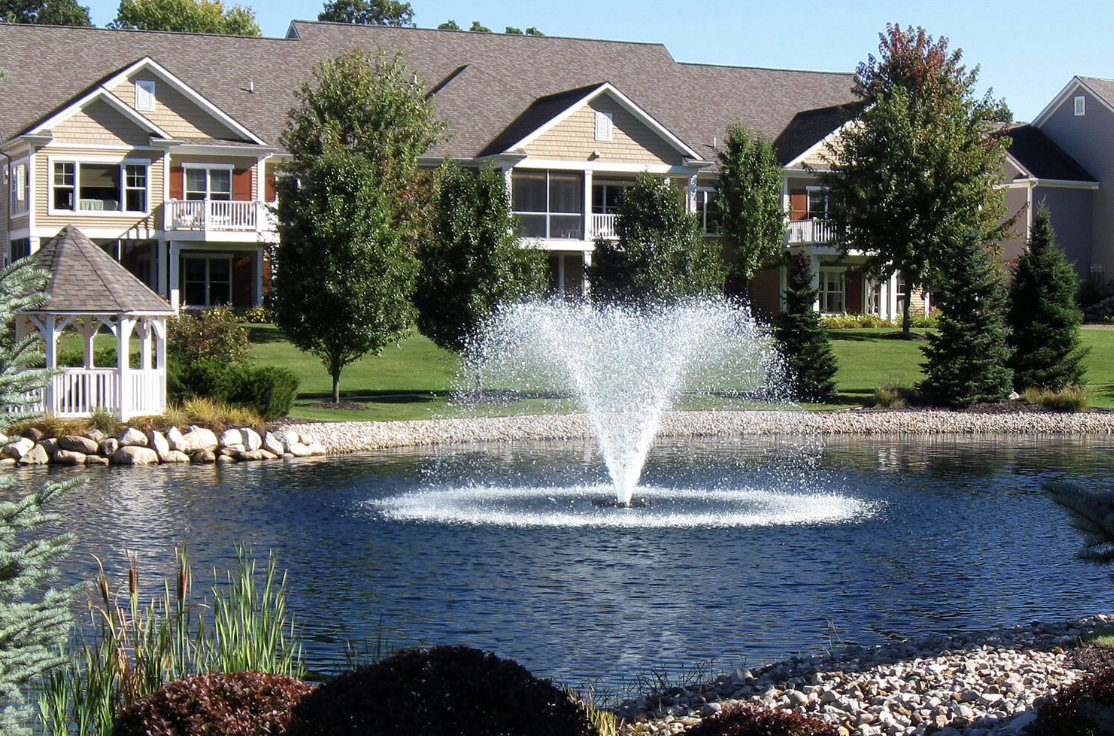 Floating aerating fountain spraying in a landscaped community pond with a stone shoreline, gazebo and townhomes behind it