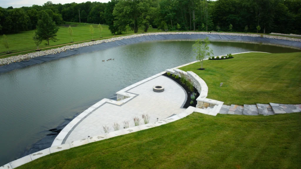 Aerial view of stone steps leading into a half-full lined pond, with a concrete and stone entertaining area, retaining walls, landscaping and geese on the water in Liberty, Missouri